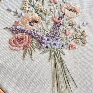 Può includere: Un cerchio da ricamo delicato che presenta un bouquet di fiori rosa, pesca e lavanda con steli verdi. I fiori sono ricamati su uno sfondo di tessuto bianco.