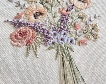 May include: A delicate embroidery hoop featuring a bouquet of pink, peach, and lavender flowers with green stems. The flowers are embroidered on a white fabric background.