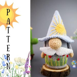 May include: Crocheted gnome amigurumi toy with a blue hat, white beard, and a yellow sun design. The gnome is standing on a wooden base with green grass and red flowers.