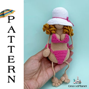May include: Crocheted doll wearing a pink bikini and a white sun hat with a pink ribbon. The doll has curly brown hair and is sitting with its legs outstretched.
