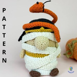 Op de afbeelding: Gehaakte kabouter verkleed als mummie met een oranje slang op zijn hoofd. De kabouter is gewikkeld in wit garen en heeft een beige gezicht en bruine voeten. Het woord "PATTERN" staat verticaal aan de linkerkant van de afbeelding.
