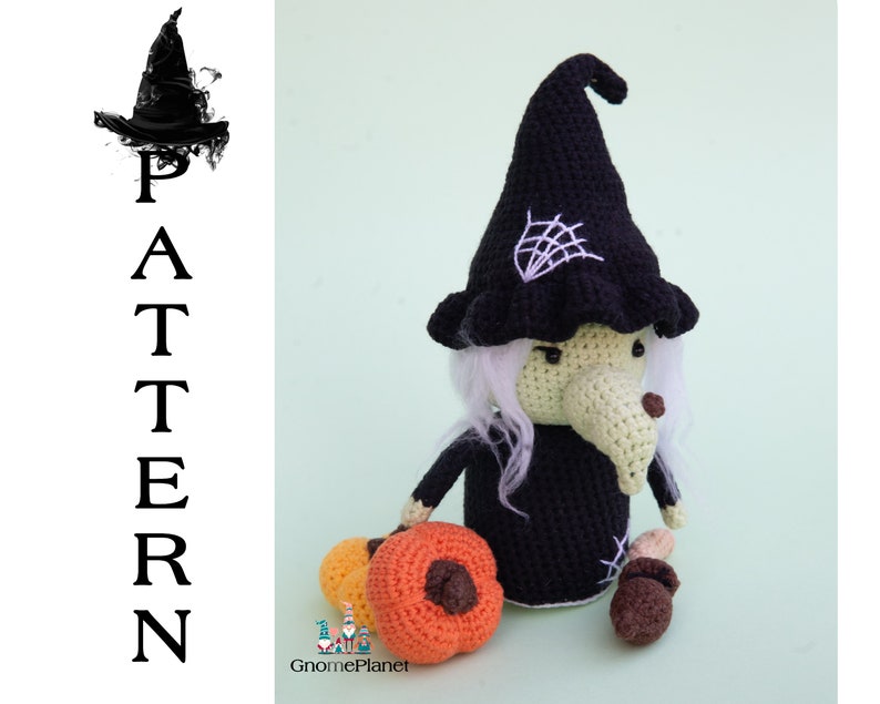 May include: Crocheted witch doll with a black hat, white hair, and a long nose. The doll is holding a small orange pumpkin and a brown acorn. The doll is sitting on a green background.