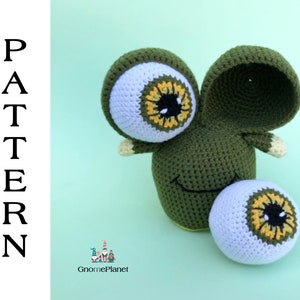 May include: Green crocheted amigurumi toy with large white eyes with yellow and orange details. The toy has a smiling mouth and a large, separate eye on the side.