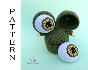 Puede incluir: Juguete amigurumi de crochet verde con grandes ojos blancos con detalles amarillos y naranjas. El juguete tiene una boca sonriente y un ojo grande separado en el lateral.
