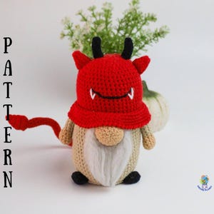 May include: A crocheted gnome wearing a red devil costume with black horns and a white beard. The gnome has a black nose and black eyes. The gnome is sitting on a white surface.