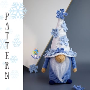 Op de afbeelding: Gehaakte blauwe gnome met een witte hoed versierd met blauwe sneeuwvlokken. De gnome heeft een witte baard en een bruine neus. Het woord "PATTERN" staat verticaal aan de linkerkant van de afbeelding.