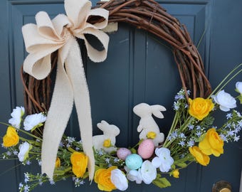 Op de afbeelding: Een bruine takkenkrans versierd met witte viltkonijnen, gele en witte bloemen en pastelkleurige paaseieren.