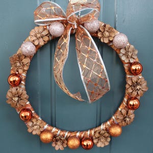 Puede incluir: Una corona decorativa con una cinta y acentos de color cobre. La corona está adornada con pequeños adornos de color cobre brillante, piñas y esferas rosas claras brillantes. La corona está diseñada para la decoración del hogar de temporada.