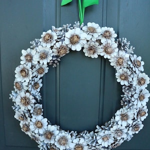 Peut inclure: Une couronne de pommes de pin blanches décorée de pommes de pin peintes en blanc et d'un ruban vert.