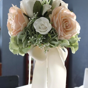 Op de afbeelding: Een bloemenboeket met grote perzikkleurige rozen, kleinere witte rozen en trossen saliegroene hortensiabloemblaadjes. Het arrangement is geaccentueerd met kleine witte vulbloemen en aan de basis vastgebonden met een lang, crèmekleurig satijnen lint.
