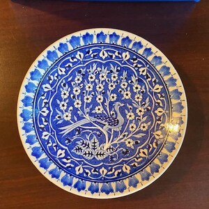 May include: A round ceramic plate featuring a blue and white pattern. The centre displays a stylised bird perched within a flowering tree. The border is decorated with a repeating petal motif. The design uses shades of cobalt blue on a white background.