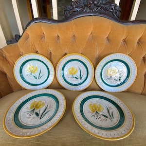 May include: A set of five ceramic plates featuring a central yellow flower illustration with green leaves. Each plate has a cream-colored center, a wide dark green band, and a yellow rim with a decorative scalloped pattern along the edge.