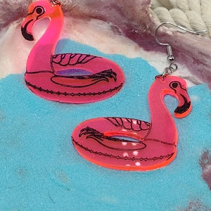 May include: A pair of pink flamingo inflatable pool float earrings. The earrings are made of clear acrylic and have a black outline. The earrings are hanging from silver hooks.