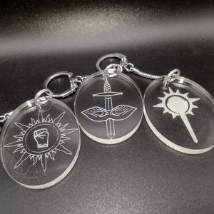 May include: Three clear acrylic keychains with different designs. The first keychain features a sun with a raised fist in the center. The second keychain features a sword and a mask. The third keychain features a star with a wand.