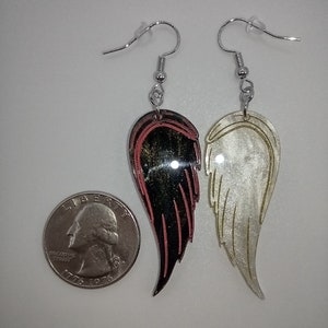 May include: A pair of dangle earrings featuring a black and red wing on one side and a white wing on the other side. The wings are made of a shiny material and have a layered look. The earrings are hanging from silver hooks.