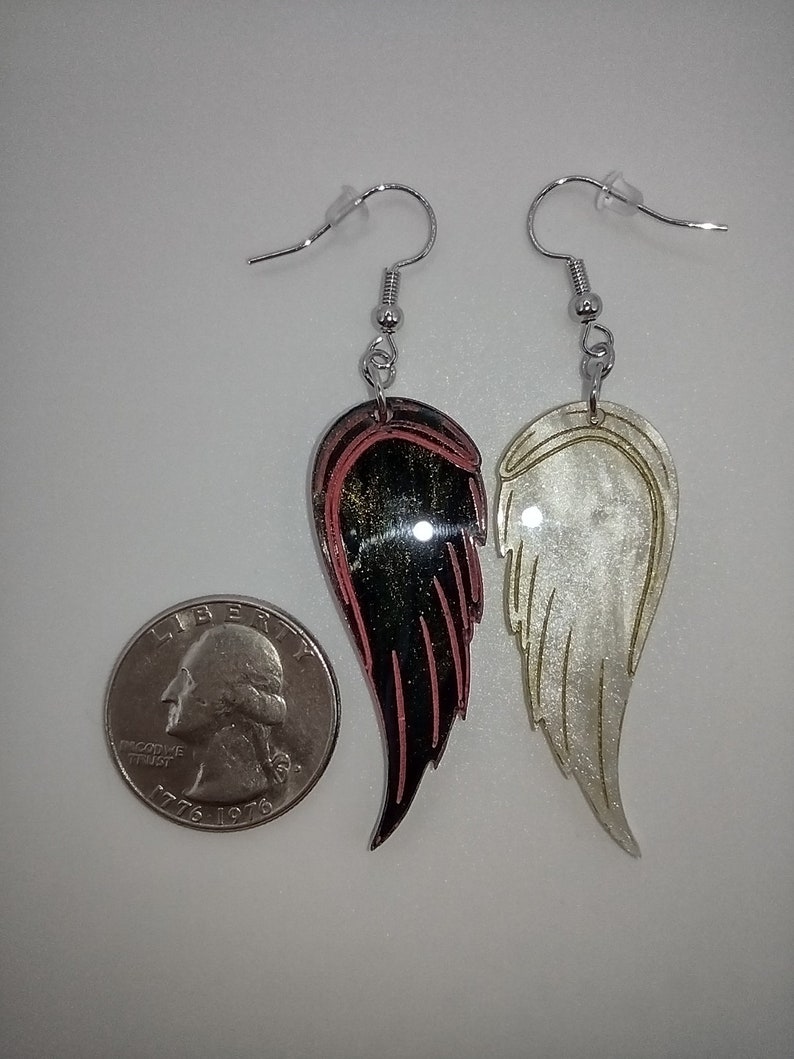 May include: A pair of dangle earrings featuring a black and red wing on one side and a white wing on the other side. The wings are made of a shiny material and have a layered look. The earrings are hanging from silver hooks.