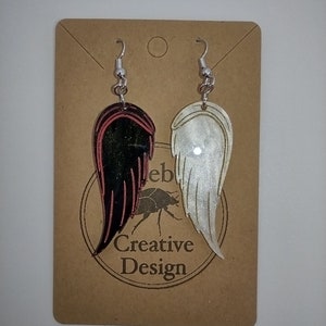 May include: A pair of dangle earrings with silver hooks. One earring features a black and red wing design, while the other earring features a white wing design. The earrings are displayed on a brown card with the text "Creative Design".