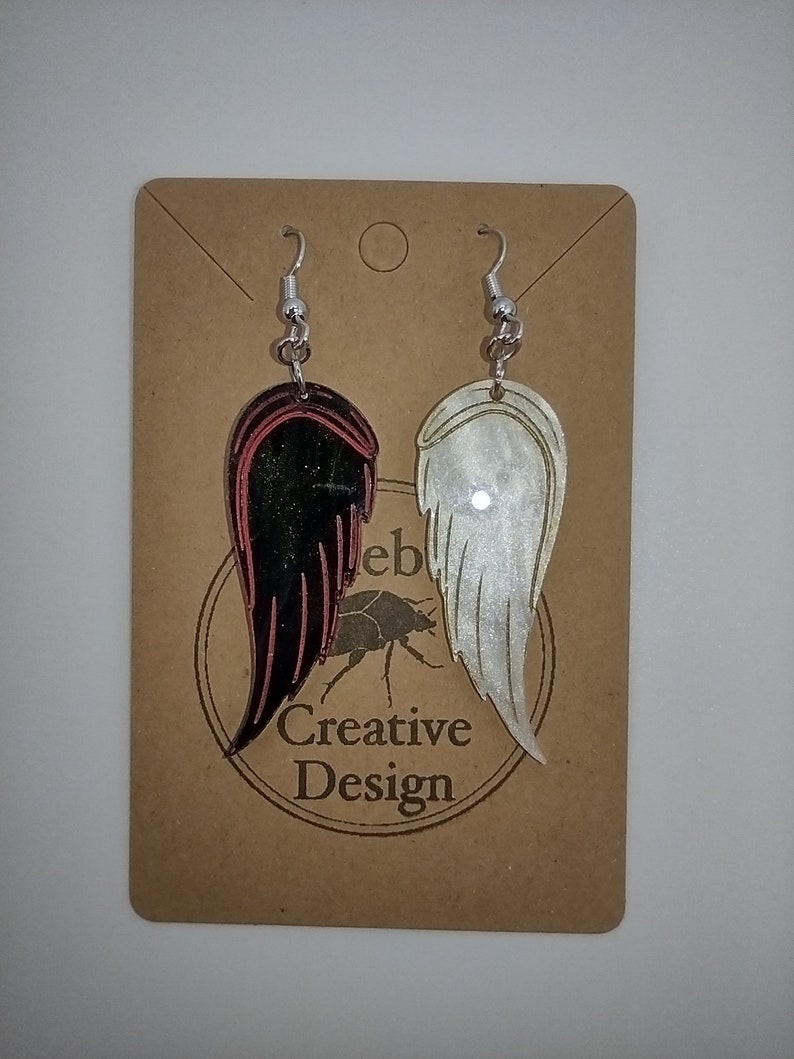 May include: A pair of dangle earrings with silver hooks. One earring features a black and red wing design, while the other earring features a white wing design. The earrings are displayed on a brown card with the text "Creative Design".