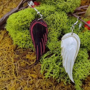 Two acrylic wing earrings. One black with red painted accents and one white with gold painted accents. Lying on a bed of moss.