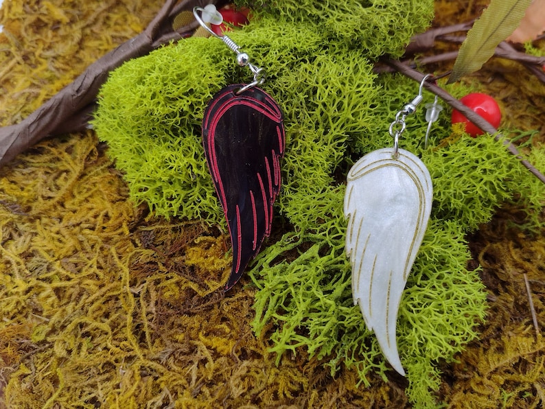 Two acrylic wing earrings. One black with red painted accents and one white with gold painted accents. Lying on a bed of moss.
