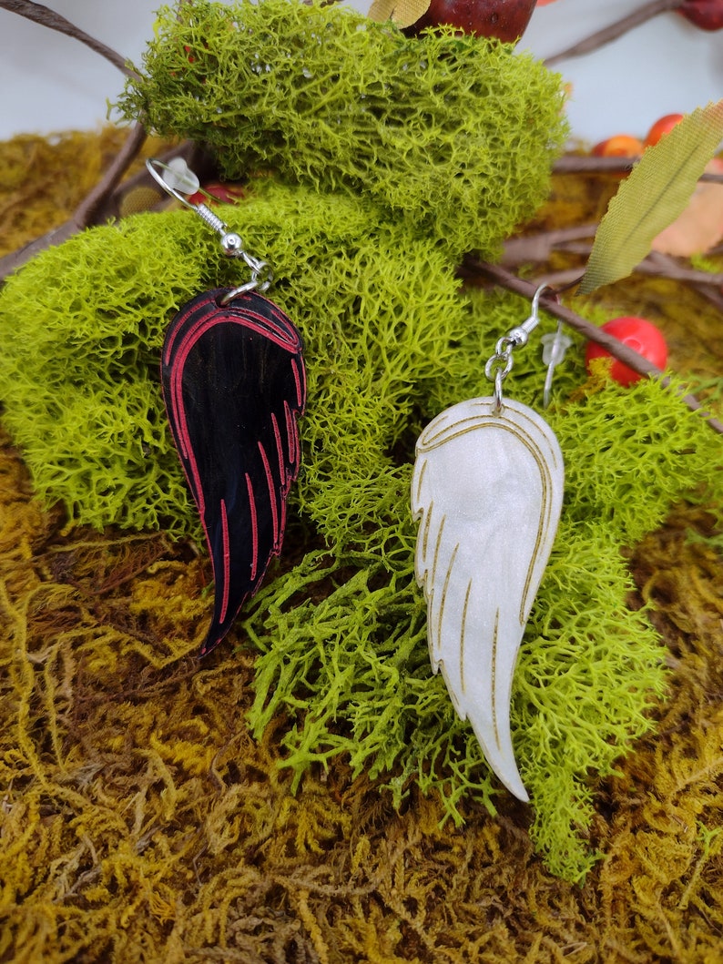 May include: A pair of dangle earrings featuring a black and red wing on one side and a white wing on the other. The wings are crafted from wood and have a painted finish.