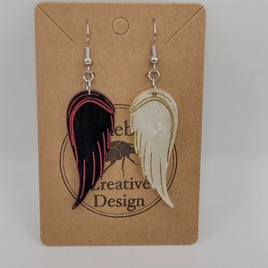 May include: A pair of dangle earrings with a black and red wing on one side and a white wing on the other side. The earrings are hanging from a brown card with the text "Creative Design" on it.