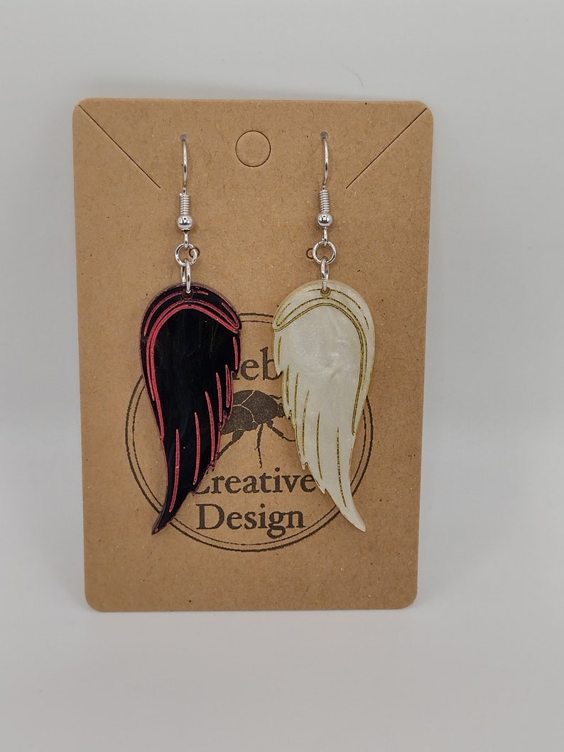 May include: A pair of dangle earrings with a black and red wing on one side and a white wing on the other side. The earrings are hanging from a brown card with the text "Creative Design" on it.