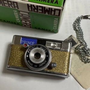 May include: A silver and gold Prince Ruby camera with a blue label that says "Prince" and a black lens with the text "Prince Ruby" and "Made in Japan".