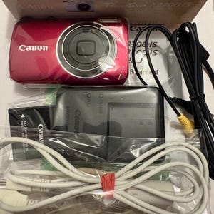 May include: A red Canon PowerShot A3300 IS digital camera with a charger, battery, and cables. The camera is in its original packaging. The kit includes a battery, charger, and various cables for connectivity.