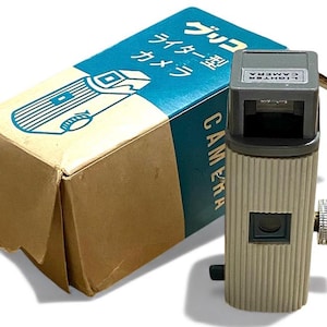 May include: A vintage camera shaped like a lighter, with a beige and gray body. The camera has a silver dial and a small lens. The box is blue and tan with Japanese text and a diagram of the camera.