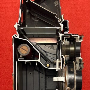 Könnte beinhalten: Eine Schnittansicht einer Vintage-Kamera, die die inneren Funktionsweisen des Mechanismus enthüllt. Die Kamera ist aus Metall und hat eine schwarze Oberfläche. Das Innere ist freigelegt und zeigt die Zahnräder, Federn und andere Komponenten, die die Kamera zum Laufen bringen.