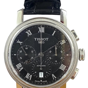 May include: A black Tissot 1853 automatic watch with a silver bezel and a black leather band. The watch face has Roman numerals and three sub-dials. The watch is Swiss made.