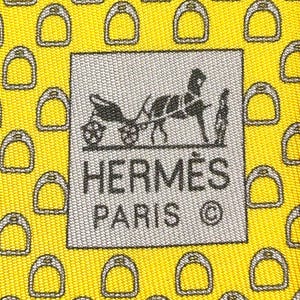 Könnte beinhalten: Eine gelbe Seidenkrawatte mit einem sich wiederholenden Muster aus grauen Steigbügeln. Die Krawatte trägt ein graues Etikett mit dem Text "HERMÈS PARIS ©".