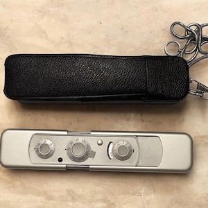 Op de afbeelding: Een zilverkleurige rechthoekige lichtmeter met drie ronde wijzerplaten met numerieke markeringen en een kleine rode stip, vergezeld van een zwarte, getextureerde lederen beschermhoes met een zilveren metalen ketting aan de zijkant.