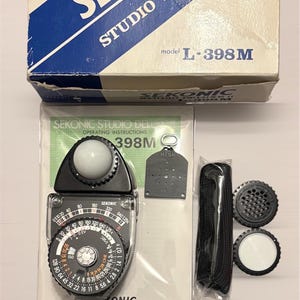 May include: A Sekonic Studio Deluxe II light meter kit. The kit includes the light meter, a black leather case, a lens cap, and operating instructions. The box is white and blue with the Sekonic logo. The model number is L-398M.