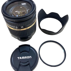 May include: A black Tamron SP 17-50mm F/2.8 lens with a lens hood, lens cap, and filter. The lens has gold lettering and markings. The lens cap has the Tamron logo in white. The lens hood is black and has a petal shape.