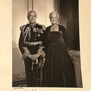 May include: A black and white photograph of a man and a woman in formal attire. The man is wearing a military uniform with medals and a sword. The woman is wearing a long black dress with a jeweled necklace. The photo is signed "George I.  1967" and "Pauline Deimer".