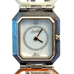 May include: A silver-toned Jean Cocteau Swiss Made watch with a rectangular face and a light-colored dial. The watch features a silver-toned band with a textured design and a clasp. The watch hands are black.