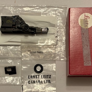 May include: A black metal camera component marked with the number 822, a small black rectangular piece, and a black rubber O-ring. These items are contained in clear plastic bags next to a red cardboard box featuring the Leitz logo and text.