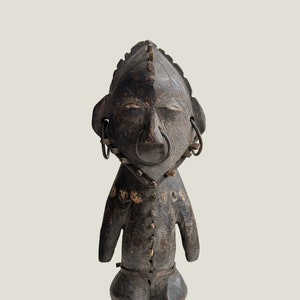 Peut inclure: Une figurine en bois brun foncé représentant une personne avec un visage stylisé et de grandes oreilles. La figurine a un design simple et géométrique et est décorée d'anneaux et de clous en métal.