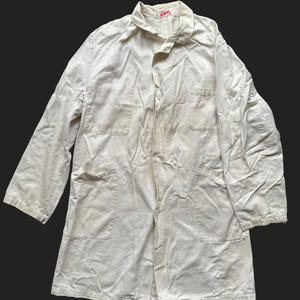 May include: A white work coat with a red "ROGER" label on the collar. The coat has two large pockets on the front and a button closure.