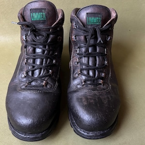 Pode incluir: Um par de botas de caminhada em couro preto, desgastadas. As botas têm atacadores pretos, ilhós de metal e o nome da marca "LIMMER" em letras verdes na língua. As solas são grossas e resistentes, adequadas para atividades ao ar livre.