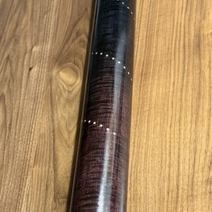 May include: A cylindrical wooden rolling pin with a dark purple and black stained finish featuring a visible wood grain pattern. The surface is decorated with a series of small, white circular inlays arranged in a spiraling pattern along the length.