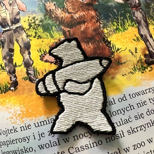 May include: White embroidered patch of a bear with black outline. The bear is standing on its hind legs with its arms outstretched. The patch is on a page of a book with text about a bear named Wojtek.
