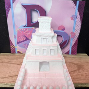May include: A white and pink miniature temple with a small tower on top. The temple has a doorway and windows. The base of the temple has a row of small white figures.