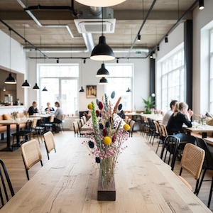 以下が含まれることがあります： 長テーブルのあるレストランのインテリア。長方形の木製花瓶にカラフルな花の装飾が施されています。黒いペンダントライトが天井から吊り下げられ、窓の近くのテーブルに人々が座っています。