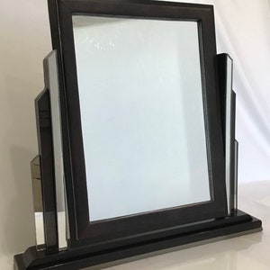 May include: A black framed vanity mirror with a rectangular shape. The mirror has a dark frame and is supported by a base with mirrored accents on each side. The design is Art Deco inspired.