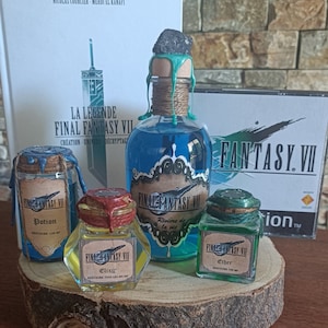 Puede incluir: Cuatro botellas de vidrio con líquido verde, azul y amarillo, etiquetadas "Final Fantasy VII" con diferentes nombres de pociones. Las botellas están dispuestas sobre una rodaja de madera con una botella azul más grande en el centro. Las botellas tienen tapones de corcho con sellos de cera. Un libro y una caja de juego son visibles en el fondo.