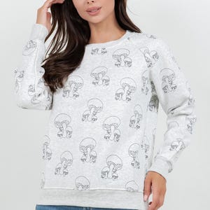 May include: A light gray sweatshirt with a repeating black outline mushroom pattern. The long-sleeved crew neck top is made of a soft material and is paired with blue jeans. The sweatshirt has a ribbed hem and cuffs.