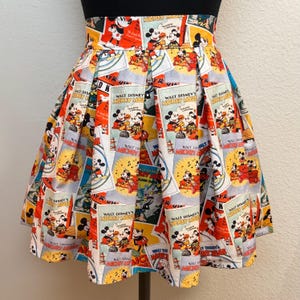 Può includere: Una gonna plissettata bianca con una stampa colorata di copertine di fumetti vintage di Topolino. La gonna è di colore rosso, giallo, blu e nero.